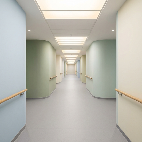 Ein heller, endloser Flur in einer modernen Pflegeeinrichtung ohne sichtbare Türen am Ende. Der Boden ist matt und kontrastarm, die Wände sind in sanften Pastellfarben gestrichen. Ein biodynamisches Lichtsystem an der Decke simuliert Tageslicht. Keine Personen im Bild.