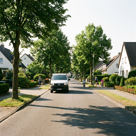 Ein weißer Kleinbus fährt durch ein gepflegtes Wohngebiet mit grünen Bäumen und Einfamilienhäusern an einem sonnigen Morgen. Realistische Szene, keine Schriftzüge.