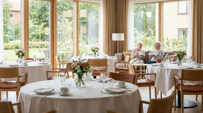 Ein moderner, heller Speisesaal in einer gepflegten Seniorenresidenz mit ansprechend gedeckten Tischen, weißen Tischdecken und frischen Blumen. Im Hintergrund unterhalten sich zwei Senioren freundlich bei einer Tasse Kaffee.
