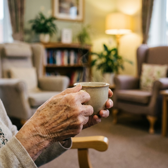 Eine Nahaufnahme von Händen einer älteren Person, die entspannt eine warme Teetasse hält. Im Hintergrund ein unscharfer, gemütlicher Ruheraum mit bequemen Sesseln in einer Pflegeeinrichtung.