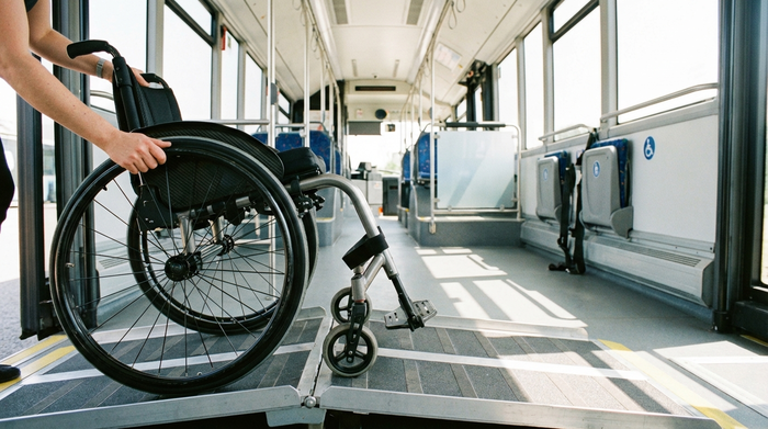 Detailaufnahme eines modernen Rollstuhls, der über eine sichere Metallrampe in den geräumigen Innenraum eines Transportbusses geschoben wird. Sauberer, gut beleuchteter Fahrzeuginnenraum.