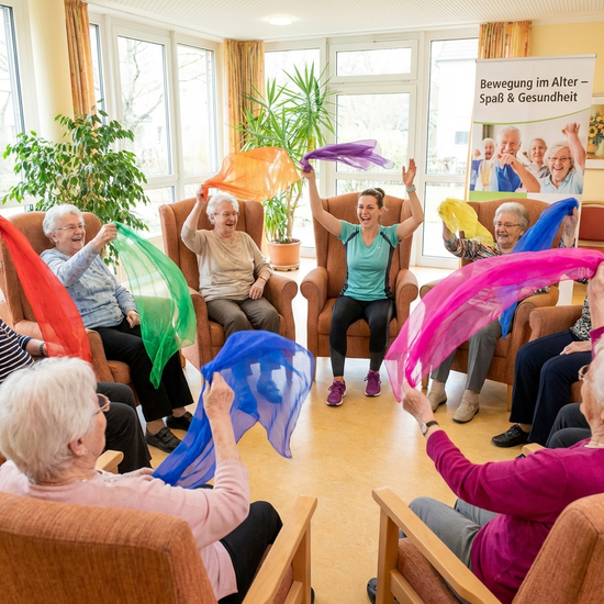 Eine Gruppe älterer Menschen sitzt im Kreis auf bequemen Stühlen und macht leichte Gymnastikübungen mit bunten Tüchern. Eine motivierte Therapeutin leitet die Gruppe freudig an.