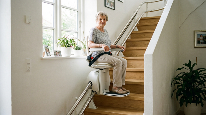 Ein moderner Treppenlift in einem gepflegten Einfamilienhaus. Eine ältere Dame fährt sicher und entspannt die Treppe hinauf. Helles Treppenhaus mit Holzstufen und weißen Wänden, realistische und sichere Umgebung.
