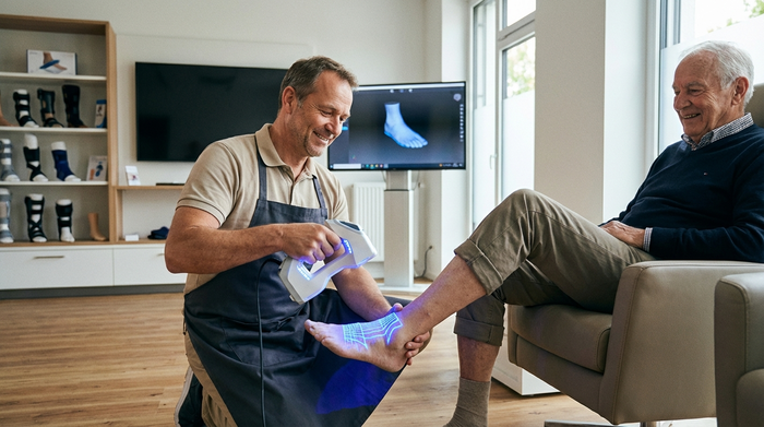 Ein Orthopädietechniker scannt den Fuß eines älteren Herrn mit einem modernen, leuchtenden 3D-Handscanner. Aufgeräumtes Sanitätshaus, professionelle und freundliche Umgebung, fotorealistisch ohne Text.
