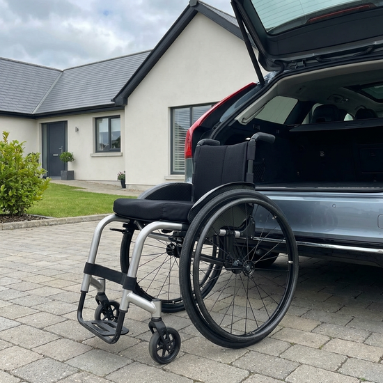 Ein moderner, leichter Aluminium-Rollstuhl steht zusammengeklappt neben dem geöffneten Kofferraum eines Autos. Ein gepflegter Vorplatz, praktische Alltagssituation, realistisches Licht, keine Beschriftungen.