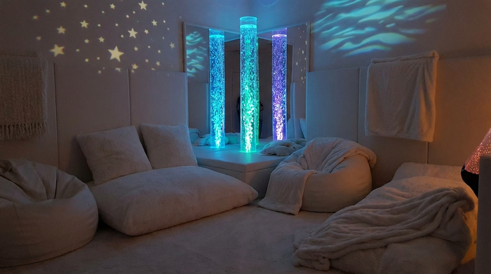Ein gemütlicher Snoezelen-Raum mit gedimmtem Licht, einer sanft blubbernden, farbig beleuchteten Wassersäule und bequemen weißen Sitzkissen auf dem Boden. Die Atmosphäre ist extrem beruhigend, weich und entspannend.