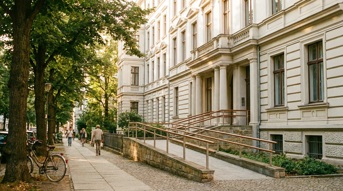 Ein Blick auf historische, gepflegte Altbauten in einer ruhigen Straße an einem sonnigen Tag, mit großen Bäumen und einem barrierefreien Zugangsweg zu einem der Häuser.