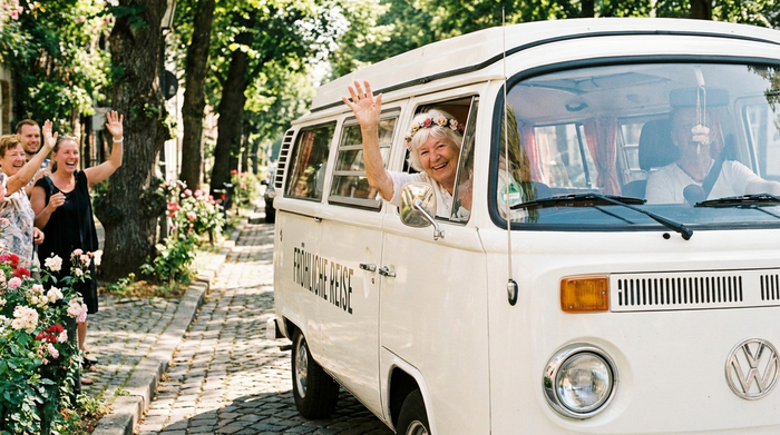 Eine Seniorin winkt fröhlich aus dem geöffneten Fenster eines weißen Kleinbusses, während das Fahrzeug in einer von Bäumen gesäumten Straße abfahrbereit steht. Fröhliche, unbeschwerte Stimmung.