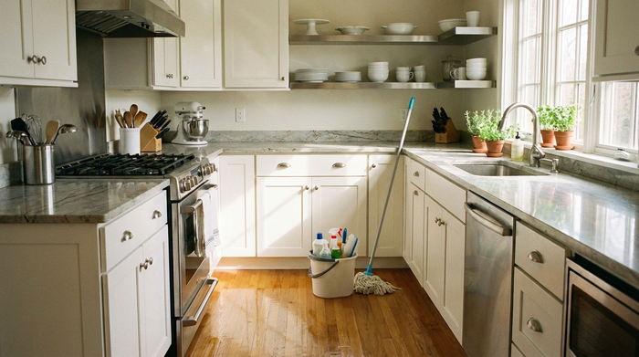 Eine ordentliche, helle Küche mit frisch gewischten Böden und sauberen Arbeitsflächen. Ein Eimer mit Reinigungsmitteln und ein Wischmopp stehen ordentlich in der Ecke. Realistische, saubere Umgebung, warmes Tageslicht, keine Schriftzüge.