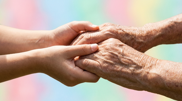 Eine emotionale Nahaufnahme von jungen Händen, die liebevoll und beruhigend die faltigen Hände einer älteren Person halten. Im unscharfen Hintergrund sind sanfte Pastellfarben zu erkennen.