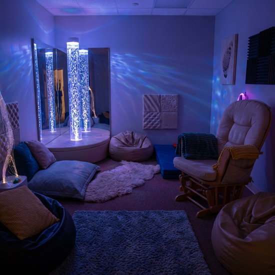 Ein gemütlicher Snoezelen-Raum in einem Pflegeheim mit gedimmtem, bläulichem Licht, einer sanft blubbernden Wassersäule und bequemen Sitzkissen. Entspannende, therapeutische und sichere Umgebung.