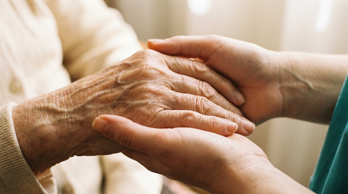 Nahaufnahme der von Falten gezeichneten Hände einer älteren Person, die friedlich in den behutsamen Händen einer jüngeren Pflegekraft ruhen. Vertrauensvolle Geste, weiches Licht, Fokus auf Geborgenheit.