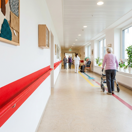 Ein breiter, heller Flur in einer Seniorenresidenz mit einem farblich deutlich abgesetzten, roten Handlauf an einer weißen Wand. Demenzgerechte, barrierefreie Gestaltung ohne Hindernisse.