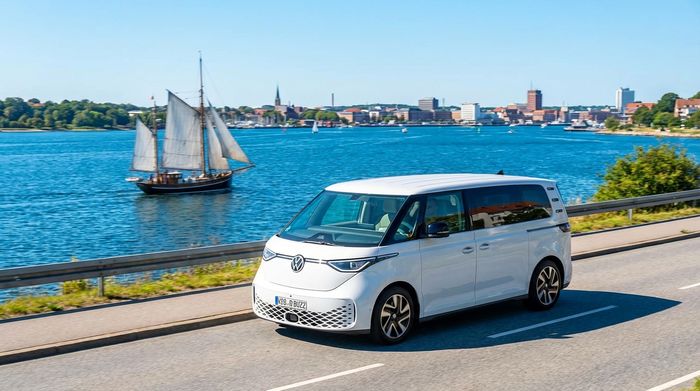 Ein moderner, weißer Kleinbus fährt an der malerischen Kieler Förde entlang. Im Hintergrund ist das strahlend blaue Wasser und ein vorbeifahrendes Segelschiff zu sehen, sonniges Wetter.