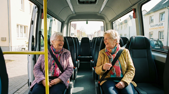 Zwei ältere Damen sitzen fröhlich plaudernd nebeneinander in den bequemen Sitzen eines Kleinbusses, sicher angeschnallt und entspannt auf dem Weg zur Tagespflege.