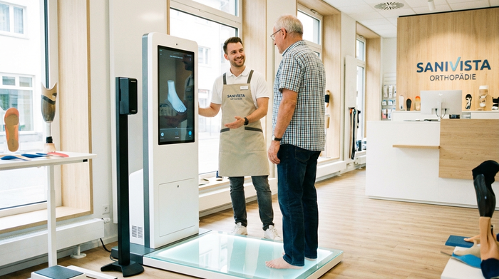 Ein freundlicher Orthopädietechniker in sauberer Arbeitskleidung steht in einem hellen, modernen Sanitätshaus neben einem digitalen 3D-Fußscanner. Ein älterer Herr steht barfuß auf der beleuchteten Glasplatte des Scanners, während der Techniker aufbauend lächelt. Die Umgebung wirkt hochtechnologisch und vertrauenswürdig.
