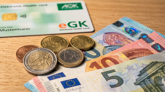 Eine detailreiche Nahaufnahme von sauberen Euro-Münzen und Geldscheinen auf einem hellen Holztisch neben einer unscharfen, stilisierten Gesundheitskarte. Warmes Licht, professionelle und realistische Fotografie.