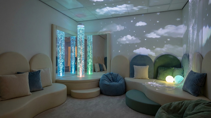 Ein atmosphärischer Snoezelen-Raum mit gedimmtem, sanftem Licht. Eine leuchtende Wassersäule mit aufsteigenden Bläschen steht im Fokus. Bequeme, weiche Liegelandschaften laden zur Entspannung ein. Beruhigende, friedliche Stimmung ohne Text.