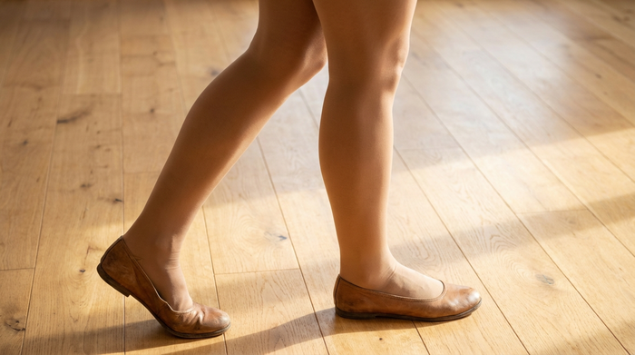 Eine Nahaufnahme von gesunden, aktiven Beinen in unauffälligen, hautfarbenen Kompressionsstrümpfen. Die Person trägt bequeme flache Schuhe und steht auf einem hellen, warmen Holzboden.
