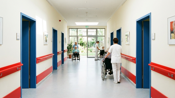 Ein breiter, heller Flur in einer Pflegeeinrichtung mit farblich abgesetzten roten Handläufen und blauen Türen zur besseren Orientierung. Das natürliche Tageslicht fällt durch große, bodentiefe Fenster am Ende des Ganges.