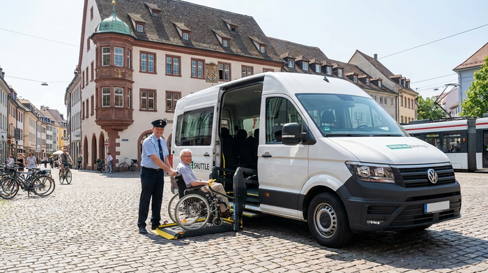 Ein moderner, rollstuhlgerechter Kleinbus parkt vor einem charmanten Freiburger Altbau. Ein freundlicher Fahrer hilft einem lächelnden Senior rücksichtsvoll beim Einsteigen. Sonniges Wetter, saubere Umgebung, realistische Straßenszene.