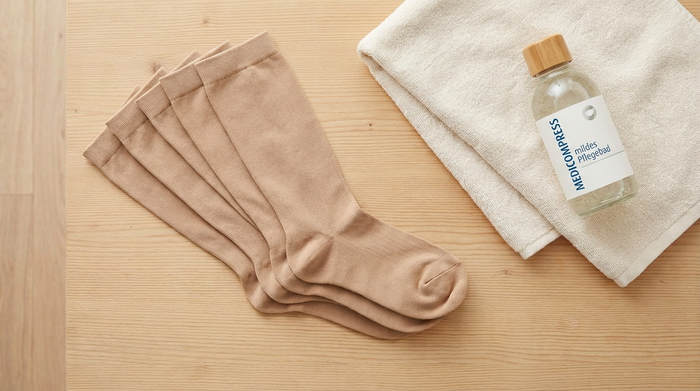Fein säuberlich auf einem hellen Holztisch ausgebreitete, frische Kompressionsstrümpfe neben einem weichen Handtuch und einem milden Pflegebad. Die Szene wirkt reinlich, aufgeräumt und hygienisch.