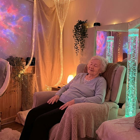 Eine ältere Frau sitzt in einem gemütlichen Snoezelen-Raum mit beruhigenden, sanften Lichteffekten und Wassersäulen. Sie wirkt entspannt und friedlich.