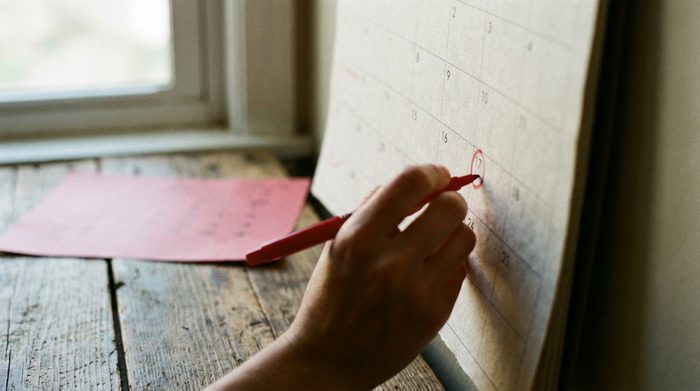 Eine Nahaufnahme eines Wandkalenders, auf dem ein bestimmtes Datum mit einem roten Stift markiert wird. Daneben liegt ein unscharfes, rosafarbenes Papier auf einem Holztisch. Natürliches Licht, realistischer Stil ohne lesbaren Text.