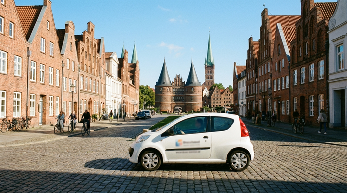 Ein kleines, weißes Auto eines Pflegedienstes fährt durch eine malerische Straße in der Lübecker Altstadt mit historischen Backsteinhäusern im Hintergrund. Sonniges Wetter, klare Umgebung, keine lesbaren Logos.