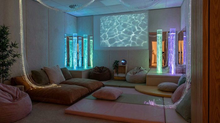 Ein friedlicher Snoezelen-Raum mit gedimmtem Licht, sanft leuchtenden Wassersäulen und bequemen Sitzkissen, der eine beruhigende Atmosphäre ausstrahlt.