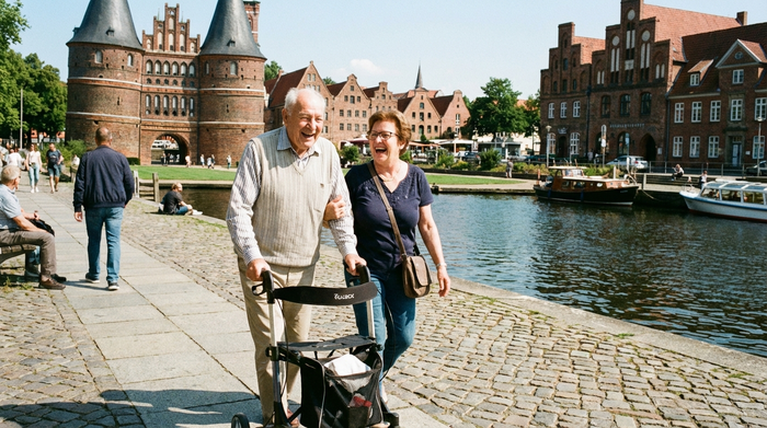 Ein älterer Herr mit Rollator und eine engagierte Alltagsbegleiterin spazieren lachend über einen gepflasterten Weg an der Trave in Lübeck. Schönes Wetter, historische Gebäude im Hintergrund, entspannte und vertrauensvolle Stimmung.
