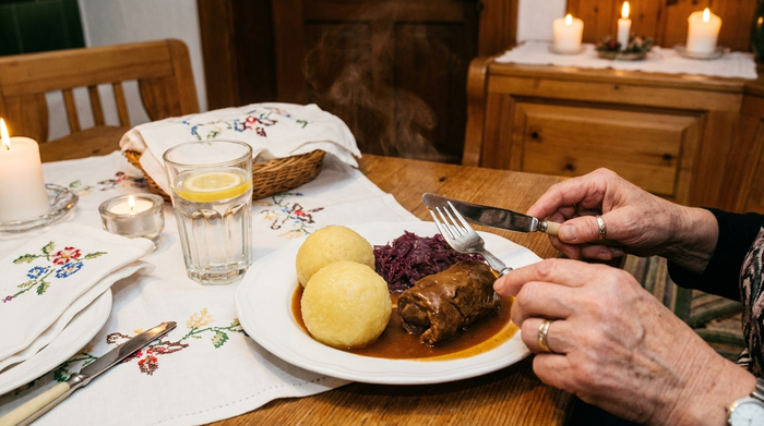 Ein liebevoll gedeckter Esstisch mit traditionellen Thüringer Klößen und Rouladen auf weißen Tellern. Ältere Hände halten Besteck. Ein Glas Wasser steht daneben. Fokus auf das appetitliche Essen, gemütliche und warme Umgebung.