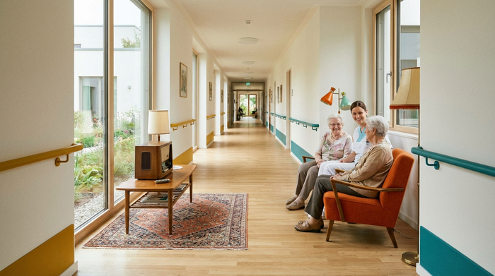 Ein heller, endloser Flur in einem modernen Pflegeheim mit farblich abgesetzten Handläufen und einer gemütlichen Sitzecke im Stil der 1960er Jahre. Ein altes Röhrenradio steht auf einem Holztischchen.