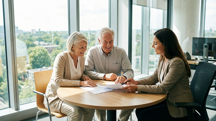 Ein älteres Ehepaar sitzt gemeinsam mit einer freundlichen Beraterin an einem runden Tisch und bespricht entspannt Dokumente. Seröse, helle Büroumgebung, keine lesbaren Texte auf den Papieren.