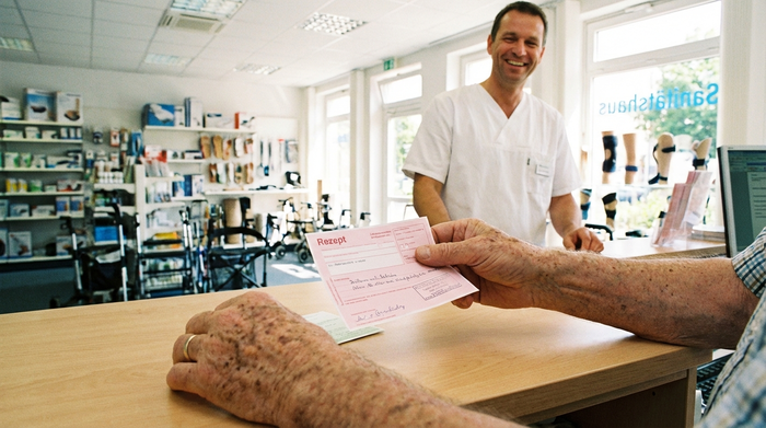 Ein detaillierter Blick auf die Hände eines älteren Herrn, der ein rosa Rezept an den Tresen eines hellen Sanitätshauses reicht. Im Hintergrund steht ein lächelnder Mitarbeiter in weißer Berufskleidung bereit.