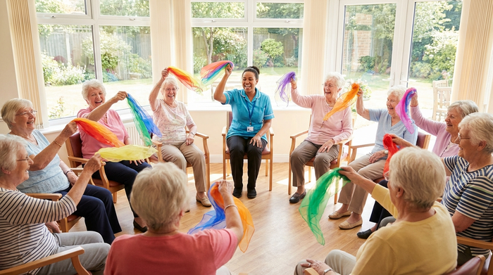 Eine Gruppe von Senioren sitzt im Kreis in einem hellen Raum und macht leichte Sitzgymnastik mit bunten Tüchern. Eine motivierende Pflegekraft leitet die Übungen an. Fröhliche Gesichter und aktive Bewegung.