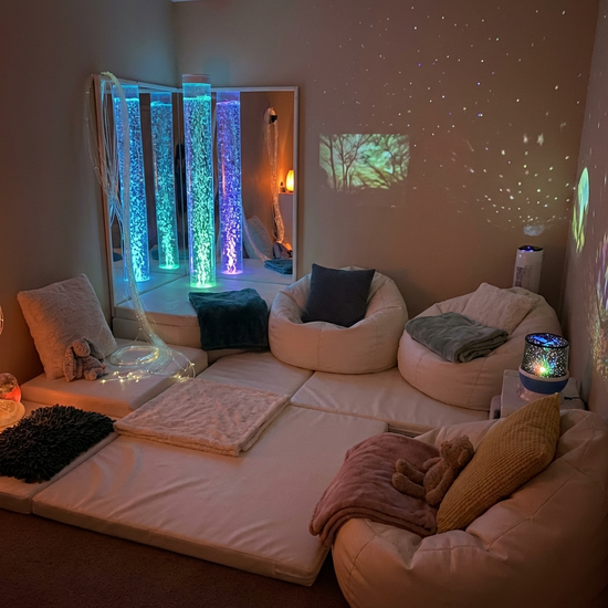 Ein gemütlicher Snoezelen-Raum mit gedimmtem Licht, einer sanft blubbernden beleuchteten Wassersäule und bequemen weißen Liegematten. Ruhige, entspannende Atmosphäre.