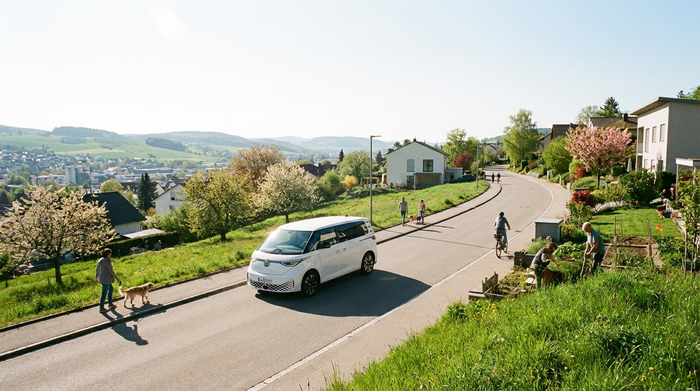 Ein moderner, weißer Kleinbus fährt eine kurvige, leicht steile Straße in einer grünen Vorstadtlandschaft hinauf. Helles Tageslicht, klare Sicht, friedliche Stimmung.