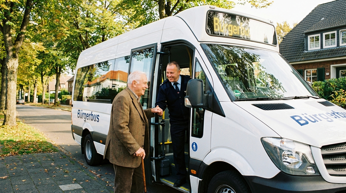 Ein freundlicher Busfahrer hilft einem lächelnden älteren Herrn mit Gehstock beim Einstieg in einen modernen, weißen Kleinbus. Der Senior trägt eine bequeme braune Jacke. Im Hintergrund sieht man eine gepflegte Wohnstraße mit Bäumen an einem sonnigen Morgen.