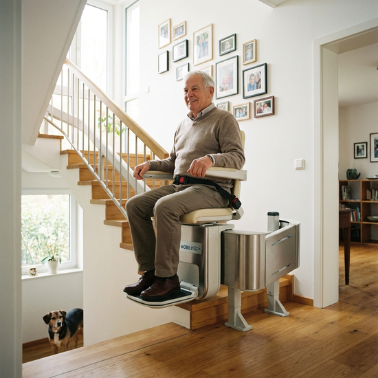 Ein moderner Treppenlift in einem gepflegten Einfamilienhaus. Ein zufriedener Senior fährt sicher die Holztreppe hinauf. Helles Treppenhaus, realistische Darstellung, Fokus auf Sicherheit und Mobilität im Alter.
