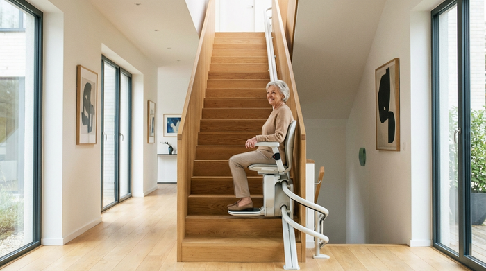 Ein moderner Treppenlift in einem gepflegten, hellen Einfamilienhaus. Eine ältere Dame fährt sicher und entspannt die Treppe hinauf, sie lächelt zufrieden. Hochwertige Holztreppe, gut beleuchteter Flur ohne störende Elemente.