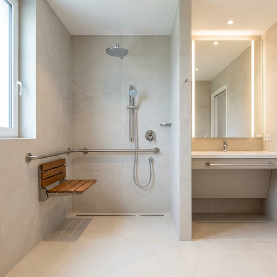 Ein modernes, hell gefliestes, barrierefreies Badezimmer mit einer geräumigen, bodengleichen Dusche. Ein stabiler Haltegriff und ein rutschfester Duschsitz sind an der Wand montiert. Sauber, sicher und elegant.