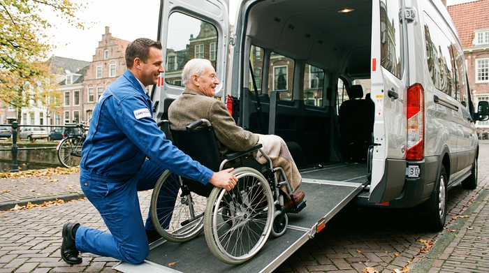 Ein professioneller Fahrer in ordentlicher blauer Dienstkleidung sichert behutsam einen älteren Herrn im Rollstuhl über eine flache Rampe im Heck eines geräumigen Transportfahrzeugs. Respektvoller Umgang, helles Tageslicht, detaillierte und realistische Darstellung.
