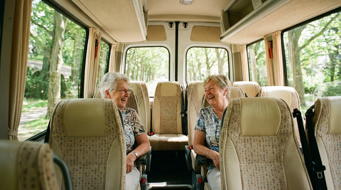 Zwei ältere Damen sitzen fröhlich plaudernd im gut beleuchteten Fahrgastraum eines Kleinbusses. Sie blicken entspannt aus dem Fenster auf eine vorbeiziehende grüne Allee. Bequeme Sitze, helle Innenausstattung, realistische und positive Stimmung.