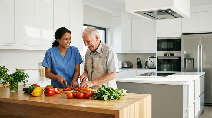 Eine engagierte Pflegekraft hilft einem älteren Herrn beim gemeinsamen Kochen in einer sauberen, modernen Küche. Frisches Gemüse liegt auf dem Tresen. Beide lachen und wirken harmonisch.