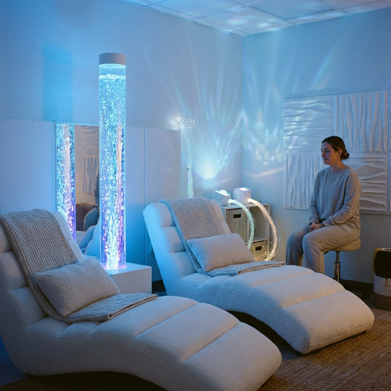 Ein gemütlicher Snoezelen-Raum mit sanftem, gedimmtem Licht in Blautönen, bequemen weißen Liegesesseln und einer beruhigend blubbernden Wassersäule. Eine Pflegekraft sitzt ruhig und aufmerksam im Hintergrund.