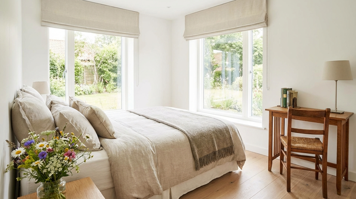 Ein helles, freundlich eingerichtetes Gästezimmer mit einem gemütlichen Bett, einem kleinen Schreibtisch und einem großen Fenster. Ein frischer Blumenstrauß steht auf dem Nachttisch. Einladende und saubere Umgebung.