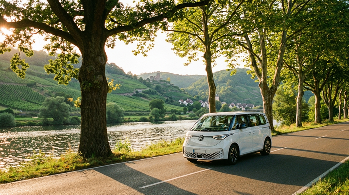 Ein moderner weißer Kleinbus fährt auf einer malerischen, von Bäumen gesäumten Straße entlang des Neckars. Im Hintergrund sind grüne Hügel und sanftes Sonnenlicht zu sehen. Ruhige und sichere Fahrszene ohne Text.