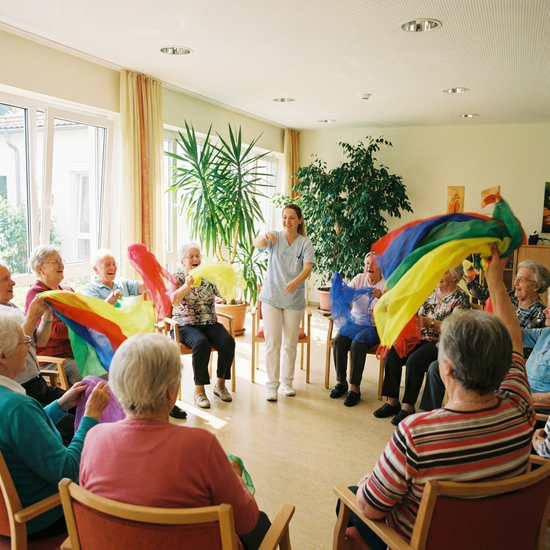 Senioren sitzen im Kreis und machen leichte Gymnastikübungen mit bunten Schwungtüchern. Eine motivierende Pflegekraft macht die Übungen vor. Helle, gut belüftete Räumlichkeiten.