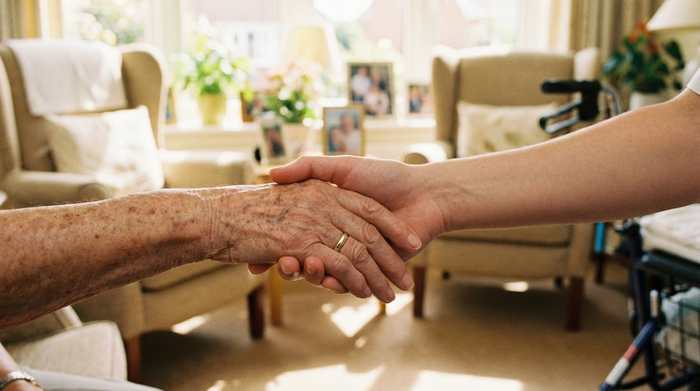 Zwei Hände, eine jung, eine alt, halten sich vertrauensvoll. Im Hintergrund unscharf ein helles, freundliches Zimmer. Symbolisiert familiäre Fürsorge und professionelle Pflege. Warmes Licht, realistisch.